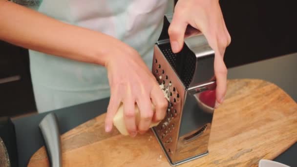 女性の手は カッティングボード上の粗い格子にチーズをこすります キッチンテーブルのミドルショット キッチンで料理する — ストック動画