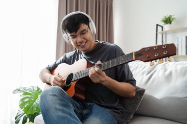 Genç yakışıklı adam akustik gitar çalıyor evdeki oturma odasında kanepede oturuyor..