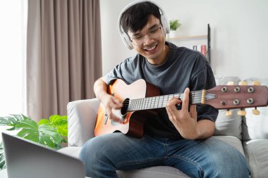 Genç yakışıklı adam akustik gitar çalıyor evdeki oturma odasında kanepede oturuyor..