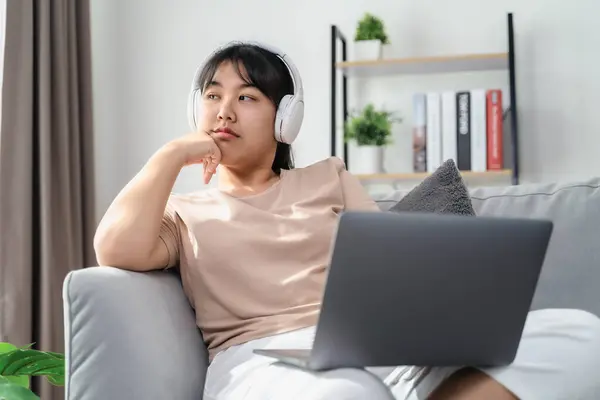 Asian woman wearing over-ear headphones feel bored while studying or working on a sofa in the living room at home.