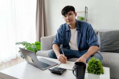  Dizüstü bilgisayar, akıllı telefon, hesap makinesi ile çalışan, evdeki oturma odasındaki kanepede oturan, ev kavramını geliştirmeye çalışan sıradan giysili adam..