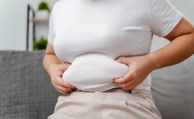 Bir kadın kanepede oturuyor ve karnını tutuyor. Kocaman bir göbeği var ve onu saklamaya çalışıyor.