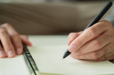 Bir adam siyah kalemle deftere yazıyor. Kişi ortaya açılan bir sayfaya yazı yazıyor. Yazı odaklanmış ve ciddi görünüyor.