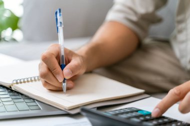 Bir adam elinde kalem ve hesap makinesiyle deftere yazı yazıyor. Kişi işine odaklanmış ve ciddi bir ruh hali içinde.