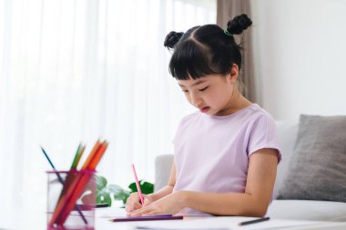 Genç bir kız kanepede oturuyor ve pastel boyalarla resim çiziyor. O, mor bir gömlek giyiyor ve saçları örgülü. Sahne huzurlu ve sakin, kız çizimine odaklanmış.