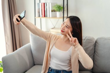 Bir kadın kanepede oturuyor ve cep telefonuyla selfie çekiyor. Gülümsüyor ve mutlu.