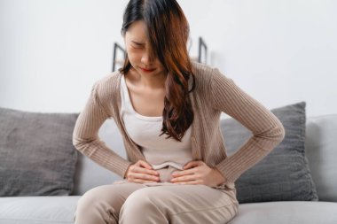 The woman with a stomachache is holding her stomach in pain, sitting on the sofa in the living room. she is suffering from a stomachache. Healthcare and medical concept.