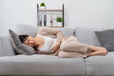 The woman with a stomachache is holding her stomach in pain, laying on the sofa in the living room. she is suffering from a stomachache. Healthcare and medical concept.