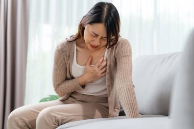 A woman is sitting on a couch with her hand holding on her chest, suffering from a heart attack. Healthcare and medical concept.