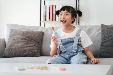 Genç bir kız kanepede ağzında lolipopla oturuyor. Gülümsüyor ve eğleniyor.