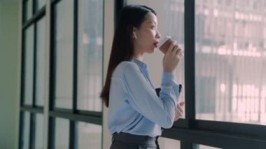 Bir iş kadını elinde bir fincan kahveyle pencereden dışarı bakıyor. O mavi bir gömlek ve siyah bir etek giyiyor.