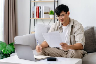 Bir adam elinde dizüstü bilgisayar ve kalemle kanepede oturuyor. Bir kağıt parçasına bakıyor ve bir şeyler yazıyor. Sahne onun bir proje üzerinde çalıştığını ya da not aldığını gösteriyor.