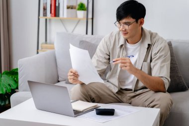 Bir adam önünde bir dizüstü bilgisayar ve bir kağıt parçasıyla bir kanepede oturuyor. Gözlük takıyor ve kağıda odaklanmış durumda.