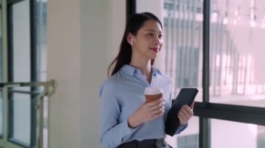 Bir kadın elinde bir fincan kahveyle koridorda yürüyor. Mavi bir gömlek giyiyor ve işine odaklanmış durumda.