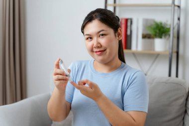 Bir kadın kanepede oturuyor ve elinde kan şekeri ölçer var. Gülümsüyor ve mutlu.