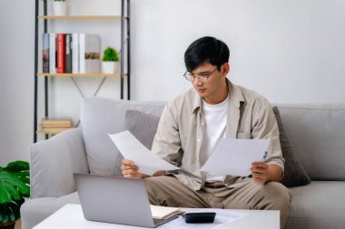 Bir adam elinde kağıtlar ve dizüstü bilgisayarla kanepede oturuyor. Gazetelere odaklanmış durumda, muhtemelen bir proje üzerinde çalışıyor ya da önemli belgeleri okuyor. Konsantrasyon ve verimlilik kavramı