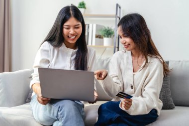 Kanepede oturan ve dizüstü bilgisayarı kullanan iki kadın. Birinin elinde kredi kartı var. Gülümsüyorlar ve bir şeyleri tartışıyor gibiler. İnternetten alışveriş yapıyorlar..