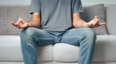 Bir adam elleri dizlerinin üzerinde bir kanepede oturuyor. Gri bir gömlek ve kot pantolon giyiyor. Rahatlama ve sakinlik kavramı, adam meditasyon yaparken ya da yoga yaparken
