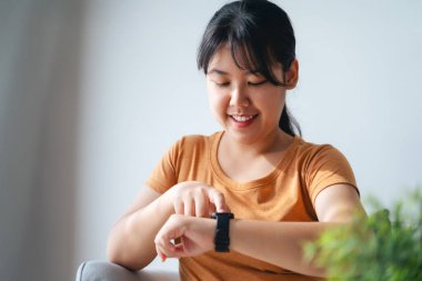 Mutlu genç kadın, bileğindeki akıllı bandı sağlık bilgilerini kontrol etmek, uyarıları kontrol etmek ve alarmı kurmak için kullanıyor. Oturma odasındaki kanepede oturuyor. Teknoloji konsepti ile sağlık hizmeti.