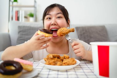 Güzel bir Asyalı kadın yüzünde bir gülümsemeyle kızarmış tavuk ve donut yiyor. Oturma odasındaki masada yiyor. Fast food, sağlıksız yemek.