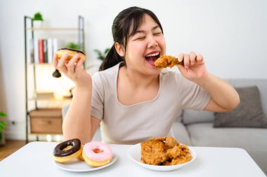 Güzel bir Asyalı kadın yüzünde bir gülümsemeyle kızarmış tavuk ve donut yiyor. Oturma odasındaki masada yiyor. Fast food, sağlıksız yemek.
