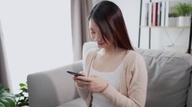 Güzel ve güzel bir Asyalı kadın, evinde bir oturma odasındaki kanepede oturan mutlu bir duyguyla akıllı bir telefon kullanıyor. Teknolojiyi ev kavramında kullanmak.