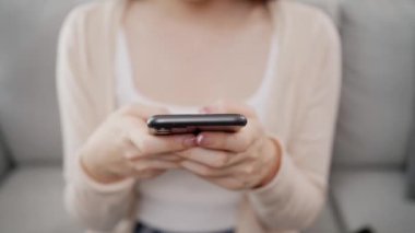 Güzel ve güzel bir Asyalı kadının akıllı telefon kullanarak evdeki oturma odasındaki bir kanepede oturduğu videoyu kapat. Teknolojiyi ev kavramında kullanmak.