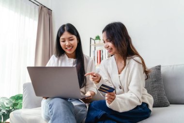 İki kadın internetten alışveriş yapmak için dizüstü bilgisayar ve kredi kartı kullanıyor. Mutlu ve neşeli hissediyorlar. Oturma odasındaki bir kanepede oturuyorlar..