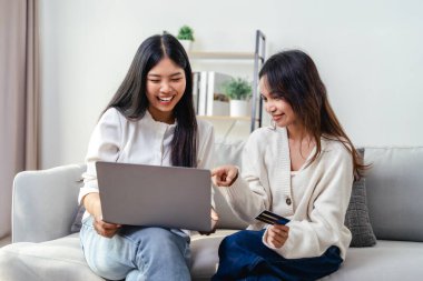İki kadın internetten alışveriş yapmak için dizüstü bilgisayar ve kredi kartı kullanıyor. Mutlu ve neşeli hissediyorlar. Oturma odasındaki bir kanepede oturuyorlar..