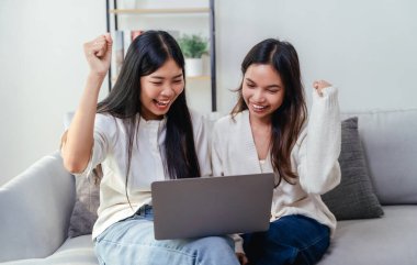 İki kadın bir dizüstü bilgisayar ekranına bakıyor ve kollarını kaldırıyor. Başarılı bir online alışverişin neşe ve mutluluk jesti olarak, oturma odasında bir kanepede oturuyorlar..