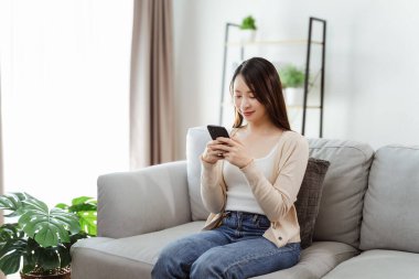 Güzel bir genç kadın akıllı telefonu tutuyor ve kullanıyor, sosyal medyayı kullanıyor, online alışveriş yapıyor, oturma odasında bir kanepede oturuyor..