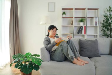 Gözlüklü Asyalı genç bir kadın oturma odasındaki kanepede kitap okuyarak mutlu bir şekilde bilgi, boş zaman, rahatlama ve hobi arıyor..