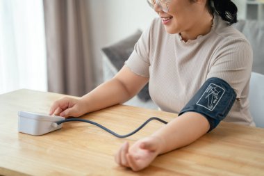 Asyalı bir kadın oturma odasında taşınabilir kan basıncı monitörü kullanıyor. Sağlık farkındalığı kavramı.