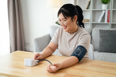 Asyalı bir kadın oturma odasında taşınabilir kan basıncı monitörü kullanıyor. Sağlık farkındalığı kavramı.