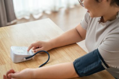 Asyalı bir kadın oturma odasında taşınabilir kan basıncı monitörü kullanıyor. Sağlık farkındalığı kavramı.