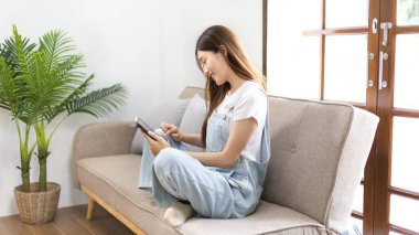 Asyalı bir kadın oturma odasındaki kanepede tabletle oturuyor, hafta sonu aktiviteleri, tatilde mutluluk, evin en sevdiği köşesi, tatlı tatlı gülümseyen güzel bir kadın..