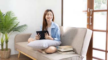 Asyalı bir kadın oturma odasındaki kanepede tabletle oturuyor, hafta sonu aktiviteleri, tatilde mutluluk, evin en sevdiği köşesi, tatlı tatlı gülümseyen güzel bir kadın..
