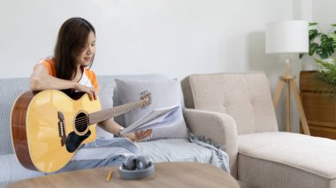 Gitar çalmayı öğrenen ya da pratik yapan bir kadın, parmaklarını gitar akorlarını tutmak için kullanıyor, notalara bilerek bakıyor, boş zamanlarından faydalanıyor, boş zamanlarında rahatlıyor..