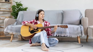 Asyalı kadın sanatçı gitar çalıyor ve salonda mutlu bir şekilde şarkı söylüyor, müzik terapisiyle rahatlıyor, müzikle boş zaman geçiriyor, müzik çalmanın keyfi, akustik gitar çalmanın keyfi,.