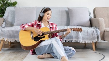 Asyalı kadın sanatçı gitar çalıyor ve salonda mutlu bir şekilde şarkı söylüyor, müzik terapisiyle rahatlıyor, müzikle boş zaman geçiriyor, müzik çalmanın keyfi, akustik gitar çalmanın keyfi,.