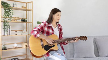 Asyalı kadın sanatçı gitar çalıyor ve salonda mutlu bir şekilde şarkı söylüyor, müzik terapisiyle rahatlıyor, müzikle boş zaman geçiriyor, müzik çalmanın keyfi, akustik gitar çalmanın keyfi,.