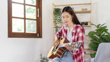 Asyalı kadın sanatçı gitar çalıyor ve salonda mutlu bir şekilde şarkı söylüyor, müzik terapisiyle rahatlıyor, müzikle boş zaman geçiriyor, müzik çalmanın keyfi, akustik gitar çalmanın keyfi,.