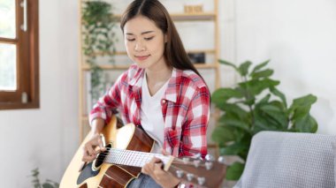 Asyalı kadın sanatçı gitar çalıyor ve salonda mutlu bir şekilde şarkı söylüyor, müzik terapisiyle rahatlıyor, müzikle boş zaman geçiriyor, müzik çalmanın keyfi, akustik gitar çalmanın keyfi,.