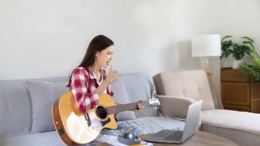 Oturma odasındaki dizüstü bilgisayarda gitar çalan ve canlı ya da canlı yayın yapan kadın sanatçı, müzik terapisiyle rahatlama, izleyicilere ya da fan kulüplerine keyif ve eğlence sunma, müzik, akustik gitar.