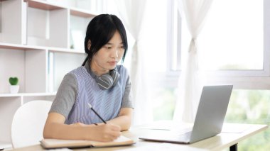 Not defterine not alan Asyalı kadın evde dizüstü bilgisayarda çalışırken, görüntülü sohbet, online iletişim, Evde kal, Yeni Normal, Mesafeli Öğrenme