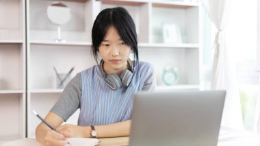 Not defterine not alan Asyalı kadın evde dizüstü bilgisayarda çalışırken, görüntülü sohbet, online iletişim, Evde kal, Yeni Normal, Mesafeli Öğrenme