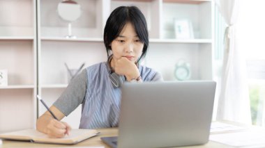 Not defterine not alan Asyalı kadın evde dizüstü bilgisayarda çalışırken, görüntülü sohbet, online iletişim, Evde kal, Yeni Normal, Mesafeli Öğrenme