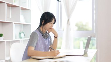Not defterine not alan Asyalı kadın evde dizüstü bilgisayarda çalışırken, görüntülü sohbet, online iletişim, Evde kal, Yeni Normal, Mesafeli Öğrenme