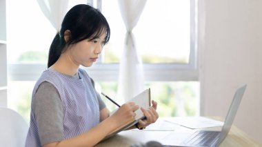 Not defterine not alan Asyalı kadın evde dizüstü bilgisayarda çalışırken, görüntülü sohbet, online iletişim, Evde kal, Yeni Normal, Mesafeli Öğrenme