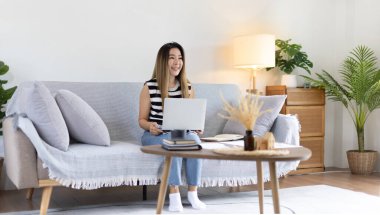 Woman using laptop to work or do homework at home with smiling face in her living room, Creating happiness at work with a smile, Live performance or video call with laptop, Work from home.